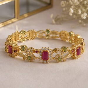 May include: A gold-toned hinged bangle bracelet featuring rectangular red stones, round green stones, and small clear pavé-set crystals arranged in an intricate metal filigree pattern throughout the circular band design.
