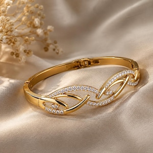 May include: A gold-toned hinged bangle bracelet featuring an openwork design of interlocking leaf-like shapes. The front section is embellished with small, clear, round-cut stones set along the curved metal edges, creating a sparkling effect.