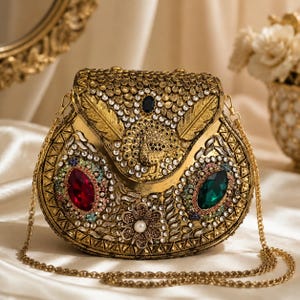 May include: A gold-toned metal clutch bag featuring an ornate peacock motif on the flap, embellished with clear crystals, a large oval red stone, a large oval green stone, and a central floral accent with a white pearl-like bead and a chain strap.