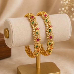 May include: Two gold-toned bangles featuring intricate metalwork, embedded with small clear stones, round red stones, and oval green stones. The bangles have a textured, filigree-style pattern throughout the band, creating a detailed and ornate appearance.