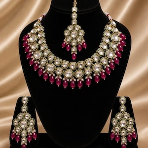 May include: A gold-coloured necklace, earrings, and headpiece set. The jewellery features clear, geometric-cut stones and ruby-coloured teardrop-shaped beads. The necklace and earrings are displayed on a black mannequin head and stand.