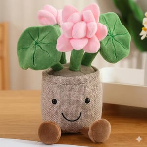 May include: A plush toy flower arrangement in a burlap pot. The pink flower has multiple layers of petals, with green leaves. The pot has a smiling face and brown feet.