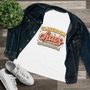 May include: White t-shirt with the text "CLASSROOM chaos COORDINATOR" in orange and yellow. The shirt is on top of a blue denim jacket, with white trainers visible at the bottom. A green plant is in the upper left corner.