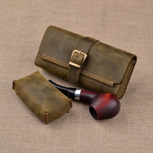 May include: A rectangular olive-green leather case with a brass buckle closure, a smaller matching leather pouch, and a tobacco pipe with a dark red bowl and a black stem. The items are constructed with visible stitching along the edges.