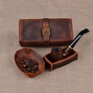 以下が含まれることがあります： 真鍮のバックルが付いた茶色のレザー製タバコポーチ、刻みタバコが入ったお揃いの茶色のレザートレイ、そして目に見えるステッチが施された小さな茶色のレザーホルダーに収められた黒いマウスピース付きの木製パイプ。