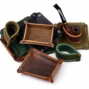 May include: A collection of leather valet trays and pipe holders in shades of brown, black, and green. The trays feature metal snap buttons at the corners and visible stitching. One tray is embossed with a small pipe icon, and a wooden pipe is included.