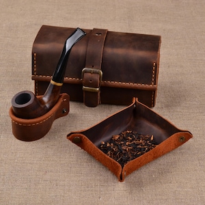 Peut inclure: Un étui à pipe en cuir marron foncé avec une fermeture à boucle, une pipe en bois avec un tuyau noir posée dans un support en cuir assorti, et un plateau carré en cuir contenant du tabac en vrac avec des coins à boutons-pression.