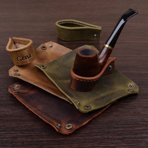 Puede incluir: Una pipa de tabaco de madera marrón oscuro con boquilla negra descansa sobre una bandeja de cuero verde con botones metálicos. Dos bandejas de cuero adicionales en marrón claro y oscuro, junto con una pequeña trabilla de cuero con el nombre Gena.