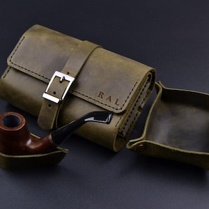 Puede incluir: Estuche de cuero verde oliva para pipa con cierre de hebilla plateada y una bandeja a juego. Una pipa marrón y negra descansa junto al estuche. Las iniciales "R.A.L." están grabadas en el estuche. El estuche está diseñado para guardar una pipa y accesorios.
