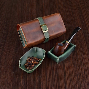 May include: A brown leather tobacco pouch with a green leather strap and brass buckle, a green leather tray containing loose tobacco, and a green leather pipe stand holding a dark brown wooden smoking pipe with a black stem.
