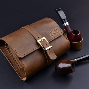 May include: A brown leather tobacco pipe case with a buckle closure, personalized with the name "Danny" and the date "5.16.26". Two tobacco pipes are next to the case. One pipe has a dark brown bowl and a black stem. The other pipe has a red and brown bowl.