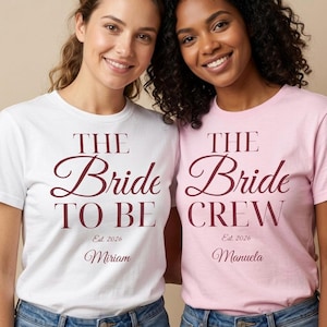 Könnte beinhalten: Ein weißes T-Shirt mit dem kastanienbraunen Text THE Bride TO BE Est. 2026 Miriam und ein hellrosa T-Shirt mit dem kastanienbraunen Text THE Bride CREW Est. 2026 Manuela. Beide T-Shirts haben kurze Ärmel und einen Rundhalsausschnitt mit Schrift.