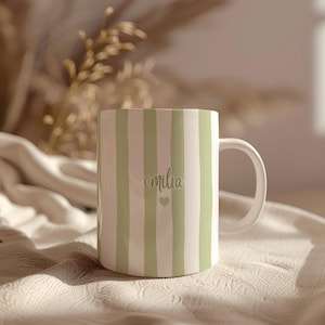 Könnte beinhalten: Eine weiße Keramiktasse mit vertikalen Streifen in einem sanften Grünton. Die Tasse trägt den Namen "emilia" in Schreibschrift sowie ein kleines Herzsymbol. Die Tasse hat einen weißen Henkel.