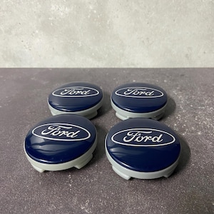 Puede incluir: Cuatro tapacubos Ford azul marino con el logotipo blanco de Ford. Las tapas tienen una base gris y un acabado brillante. Las tapas están dispuestas en forma cuadrada sobre una superficie gris.