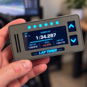 May include: A rectangular grey digital lap timer device with a black screen displaying racing data including current lap 1:34.287, best lap 1:33.912, and delta +0.375. The unit features six blue LED indicator lights, two blue arrow buttons, and text.