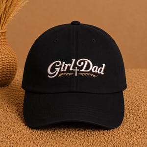 May include: A black baseball cap featuring the embroidered text "Girl Dad" in white and brown script. A small white cross symbol is positioned between the two words, with two thin, curved brown wheat-like stalks embroidered underneath the text.