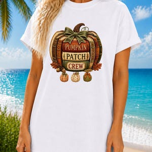 Puede incluir: Una camiseta blanca de manga corta con un gráfico de una calabaza formada por libros apilados. El diseño incluye un lazo verde en la parte superior y el texto PUMPKIN PATCH CREW en los lomos, con tres pequeñas calabazas debajo.