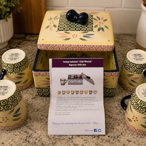 May include: A Temp-tations Old World square gift set featuring a cream-colored square box with a lid and four matching mugs. The ceramic pieces are decorated with green, pink, and blue floral patterns. A product information card is included.