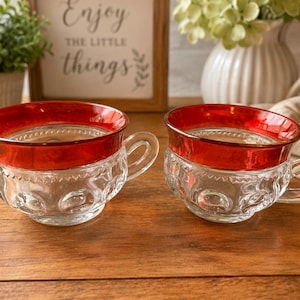 May include: Two clear glass teacups featuring a wide, metallic red band around the upper rim. The cups have a textured, embossed pattern of rounded shapes along the lower body and a small, curved handle on the side of each piece.