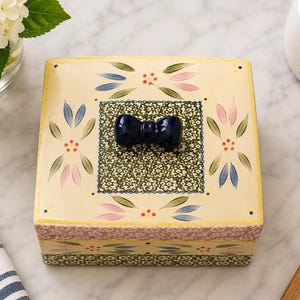May include: A square ceramic box with a cream-colored lid featuring hand-painted floral motifs in pink, blue, and green, centered with a dark blue bow-shaped handle. The sides of the box display a green and white intricate pattern with floral accents.