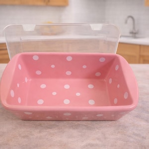 May include: A pink rectangular baking dish with white polka dots and a clear lid. The dish is sitting on a light-colored countertop. Kitchenware.