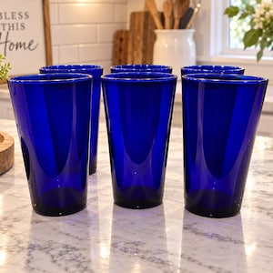 May include: A set of six cobalt blue drinking glasses with a tapered, conical shape and a smooth, transparent finish. The glasses are arranged in two rows, featuring a deep, vibrant blue color throughout the glass material with a flat base.