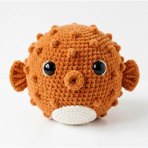 May include: A handmade, crocheted pufferfish toy in shades of orange and white. The plush toy has a round body with textured details, large black eyes, and small fins. The belly is white, and the overall design is whimsical.
