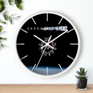 May include: A round wall clock with a white frame and a black face. The clock features the word "INTERSTELLAR" in white text, a geometric design, and white hands. The clock is approximately 12 inches in diameter.