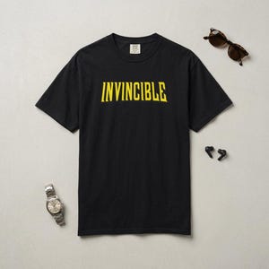 Peut inclure: T-shirt noir avec le mot "INVINCIBLE" en lettres jaunes. La chemise est accompagnée d'une montre-bracelet argentée, de lunettes de soleil marron et d'écouteurs sans fil noirs.