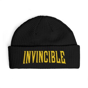 May include: Black knitted beanie hat featuring the word "INVINCIBLE" in gold lettering. The hat has a ribbed design and a folded brim. A simple, warm winter accessory.