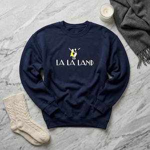 May include: Navy blue sweatshirt with the words "LA LA LAND" in white block letters. Above the text is a yellow and white graphic of two people dancing. A pair of cream-colored cable knit socks and a gray scarf are also in the image.
