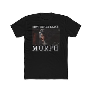 May include: Black t-shirt featuring a graphic of a person in a space helmet with the text "DON'T LET ME LEAVE" above and the name "MURPH" below. The design is in white, contrasting against the black fabric.