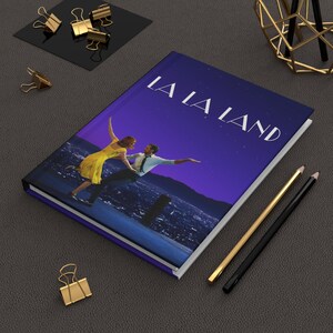 May include: A hardcover journal featuring the movie title "LA LA LAND" on the cover. The cover art depicts a couple dancing against a starry night backdrop. Gold and black pencils and gold binder clips are also visible.
