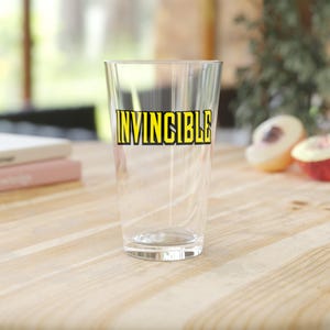 May include: A clear glass pint glass with the word "INVINCIBLE" printed in yellow with a black outline. The glass is sitting on a light wood surface. Peaches are in the background.