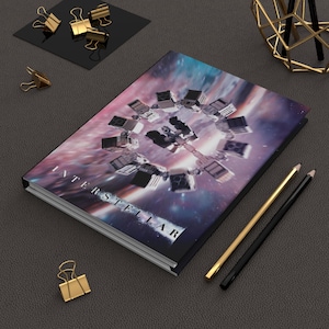 May include: A black journal with the word "INTERSTELLAR" printed on the cover. The cover features a space station design against a purple and blue galaxy background. Gold and black pencils and gold binder clips are also present.