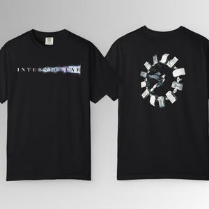 May include: Black t-shirt featuring the word "INTERSTELLAR" in white and blue lettering on the front. The back of the shirt displays a circular graphic with a space station design. The shirt is made of a soft material.