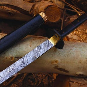Könnte beinhalten: Ein japanisch anmutendes Schwert mit gemusterter Klinge und schwarzem Griff. Die Klinge hat ein silbernes und graues Muster. Der Griff ist schwarz mit goldenen Akzenten. Das Schwert befindet sich in einer schwarzen Scheide mit goldenen Verzierungen.