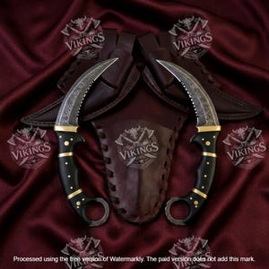 May include: Two curved karambit knives with Damascus steel blades, black handles with brass pins, and finger rings at the base. Each knife includes a brown leather sheath, with a central leather piece featuring the EB VIKINGS logo.