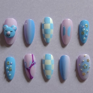 May include: A set of ten almond-shaped press-on nails featuring a pastel color palette of light blue and lavender. Designs include a 3D blue bear face, a blue and white checkerboard pattern, a BTS logo, gold star and moon charms, and pearls.