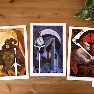 May include: Three fantasy art prints with white borders, displayed on wood. One shows a knight, another a figure with an owl, and the third a demon with a scythe. The prints feature detailed illustrations with a fantasy theme.
