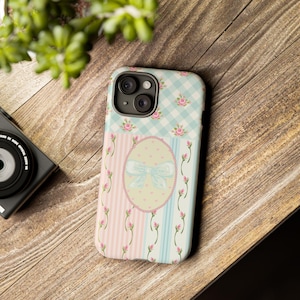 Pode incluir: Uma capa de smartphone com uma secção superior de padrão xadrez vichy azul claro e branco adornada com pequenas rosas cor-de-rosa. A parte inferior apresenta riscas verticais cor-de-rosa e azuis com botões de rosa e um laço central.