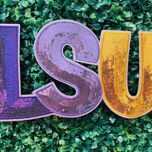 May include: Large sequined letters spelling "LSU" in purple and gold. The letters are outlined in a darker shade of their respective colors. The background is a dense arrangement of green leaves.