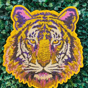 May include: A sequined tiger face patch with yellow, purple, and pink details. The tiger's face is centered, with a yellow border. The sequins create a textured, shimmering effect, perfect for adding flair to clothing or accessories.