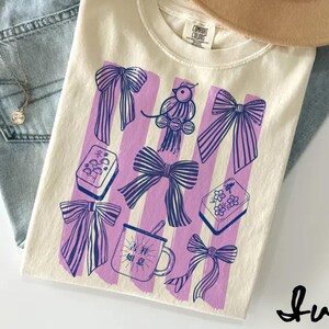 Könnte beinhalten: Elfenbeinfarbenes T-Shirt mit einem grafischen Design mit lila Schleifen, einem Vogel und anderen dekorativen Elementen. Das Shirt wird mit hellblauen Jeans und einem beigefarbenen Hut kombiniert. Das Wort "Ivory" ist in einer Schreibschrift geschrieben.