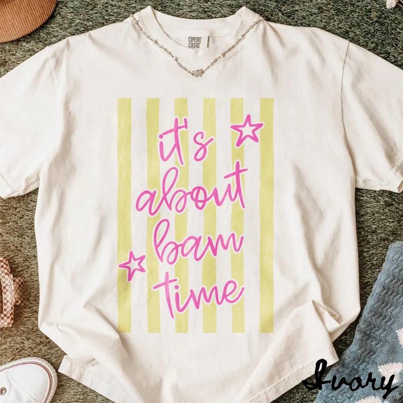 May include: Ivory t-shirt with the pink text "It's about bam time", pink stars, and yellow vertical stripes. The shirt has a Comfort Colors label and a silver necklace. The word "Ivory" is printed at the bottom right.