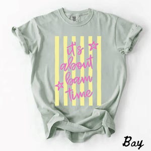 May include: A pale green t-shirt featuring the pink text "It's about bam time" with star accents, set against yellow vertical stripes. The word "Bay" is printed in the bottom right corner of the shirt.