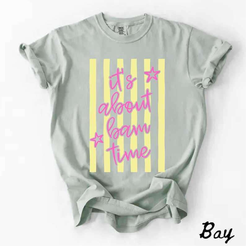 May include: A pale green t-shirt featuring the pink text "It's about bam time" with star accents, set against yellow vertical stripes. The word "Bay" is printed in the bottom right corner of the shirt.