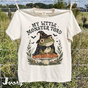 May include: An ivory short-sleeve t-shirt featuring a graphic print of a toad wearing a witch hat and cape, sitting on a mushroom. The text "MY LITTLE MONSTER TOAD" is printed above the toad, surrounded by small bats, stars, and foliage illustrations.