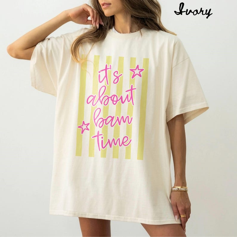 May include: Ivory t-shirt featuring vertical yellow stripes and the pink text "It's about bam time". The top has a loose fit and short sleeves, suitable for everyday wear.