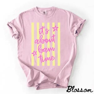 May include: A pale pink t-shirt featuring yellow vertical stripes and the pink text "It's about bam time". Two pink stars are positioned near the text. The word "Blossom" is in black script at the bottom right.